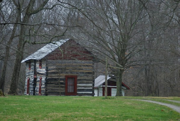 A rustic, weathered log cabin with red-framed windows stands amid a sparse forest of leafless trees. The cabin has a gabled roof made of metal that appears aged and fits the era of the building. A smaller structure is attached to the cabin's side. The surrounding ground is covered with patchy green grass, while bare trees dominate the background, creating a sense of stillness.