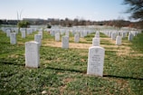 A cemetery with numerous white headstones neatly aligned in rows on a grassy field. The headstones bear inscriptions commemorating deceased military personnel. Trees are scattered in the background, and a clear blue sky is overhead. A notable monument structure is visible on the far left.