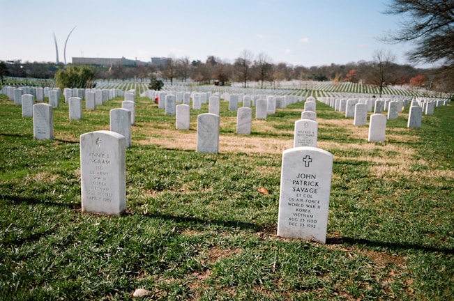 A cemetery with numerous white headstones neatly aligned in rows on a grassy field. The headstones bear inscriptions commemorating deceased military personnel. Trees are scattered in the background, and a clear blue sky is overhead. A notable monument structure is visible on the far left.
