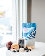 A blue bag of coffee with the branding clearly displayed is placed on a white table near a window. In front of the bag is a glass of black coffee surrounded by sliced apples, a whole apple, blackberries, and a small pile of cream. Soft natural light is coming through the window, creating a calm and cozy atmosphere.