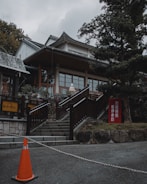A traditional architectural building with a sloping roof is situated among trees. Stone stairs lead up to the entrance, with a sculpture visible near the top. An orange traffic cone marks a barrier at the bottom of the stairs, accompanied by signs with Japanese writing.