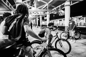 Several people with bicycles gather in what appears to be a train station. One person leans on their bike, while others stand nearby. The setting is an open area with metal beams and pillars, and the atmosphere is casual and relaxed.