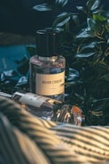 A bottle of perfume labeled 'Rose Cerise' is placed beside a variety of green foliage on a surface that appears to be a striped fabric. There is another perfume bottle lying down next to it, partially in view.