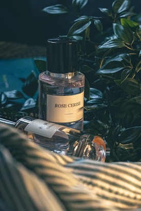 A bottle of perfume labeled 'Rose Cerise' is placed beside a variety of green foliage on a surface that appears to be a striped fabric. There is another perfume bottle lying down next to it, partially in view.