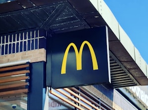 A prominent yellow McDonald's logo is displayed on a black sign attached to the exterior of a building. The structure features brickwork and wooden elements, with a clear blue sky in the background.