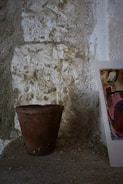 A terracotta flower pot sits on a textured stone surface with rough walls in the background. Nearby, there is a partial view of a colorful abstract painting leaning against the wall.