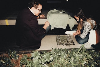 Two individuals are engaged in a game of chess on a board set up outdoors at night. One person, wearing a jacket, is seated on the ground, deeply focused on the board. The other, dressed casually, is also engrossed in the game, positioned opposite. Surrounding greenery can be seen in the foreground, providing a natural setting for this intense match.