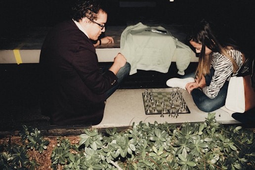 Two individuals are engaged in a game of chess on a board set up outdoors at night. One person, wearing a jacket, is seated on the ground, deeply focused on the board. The other, dressed casually, is also engrossed in the game, positioned opposite. Surrounding greenery can be seen in the foreground, providing a natural setting for this intense match.