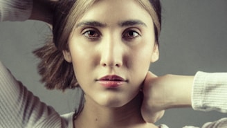 A close-up portrait of a person with a contemplative expression. The individual has light skin, dark hair, and is gazing directly at the camera. Their hands are positioned behind their neck, and they are wearing a light-colored top. The background is plain and neutral.