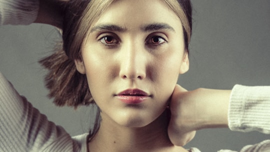 A close-up portrait of a person with a contemplative expression. The individual has light skin, dark hair, and is gazing directly at the camera. Their hands are positioned behind their neck, and they are wearing a light-colored top. The background is plain and neutral.