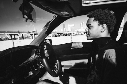A man is driving a car with a cityscape visible in the background. The image is in black and white, capturing shadows and light contrasts. The man's expression is calm and focused on the road.