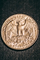 An American quarter dollar coin featuring an eagle with outstretched wings and the inscription 'UNITED STATES OF AMERICA' and 'QUARTER DOLLAR'. The text 'E PLURIBUS UNUM' is also visible above the eagle. The coin is set against a dark textured background.