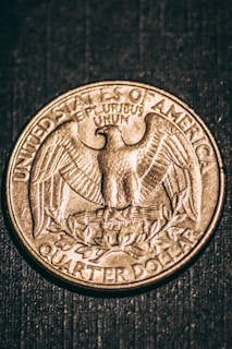 An American quarter dollar coin featuring an eagle with outstretched wings and the inscription 'UNITED STATES OF AMERICA' and 'QUARTER DOLLAR'. The text 'E PLURIBUS UNUM' is also visible above the eagle. The coin is set against a dark textured background.