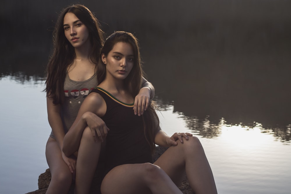 two women sitting on a rock next to a body of water