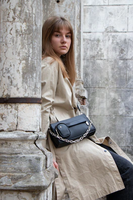 Side profile of a woman in a flowing trench coat standing against a pale stone wall.