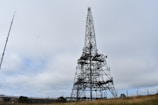 A large metal telecommunications tower stands tall against a cloudy sky. The structure is composed of intricate metalwork and is surrounded by a fenced grassy area. A smaller antenna is visible to the left, suggesting a communications site.