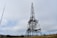 A large metal telecommunications tower stands tall against a cloudy sky. The structure is composed of intricate metalwork and is surrounded by a fenced grassy area. A smaller antenna is visible to the left, suggesting a communications site.