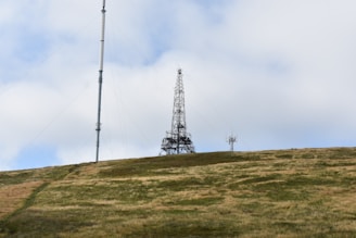 A tall radio or communication tower stands on a grassy hill under a mostly cloudy sky. The metal structure is connected by wires and is accompanied by a smaller antenna on the right.