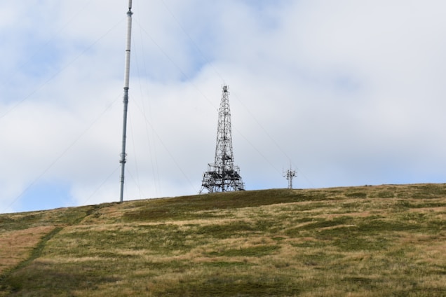 A tall radio or communication tower stands on a grassy hill under a mostly cloudy sky. The metal structure is connected by wires and is accompanied by a smaller antenna on the right.