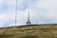 A tall radio or communication tower stands on a grassy hill under a mostly cloudy sky. The metal structure is connected by wires and is accompanied by a smaller antenna on the right.