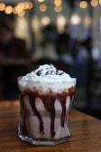 A glass filled with a creamy chocolate beverage, topped with whipped cream and drizzled with chocolate sauce. The background is softly blurred, with warm, bokeh lights creating a cozy atmosphere.