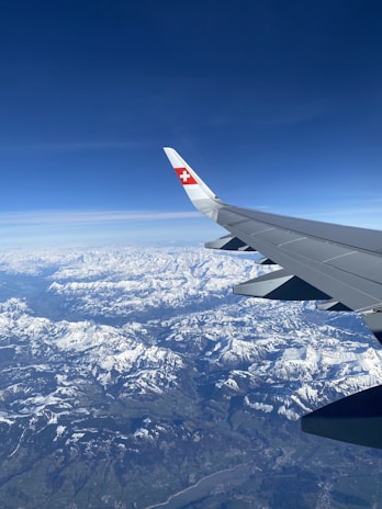 An airplane wing with a red and white Swiss flag emblem is visible against a vast, clear blue sky. Below, an impressive expanse of snow-covered mountains stretches out, offering a stunning aerial view of the landscape.