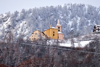 A picturesque village nestled in a snowy mountainous landscape. A prominent, rustic church with a steeple and warm yellow tones stands out against the backdrop of snow-covered trees and rugged hills.