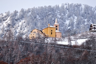A picturesque village nestled in a snowy mountainous landscape. A prominent, rustic church with a steeple and warm yellow tones stands out against the backdrop of snow-covered trees and rugged hills.