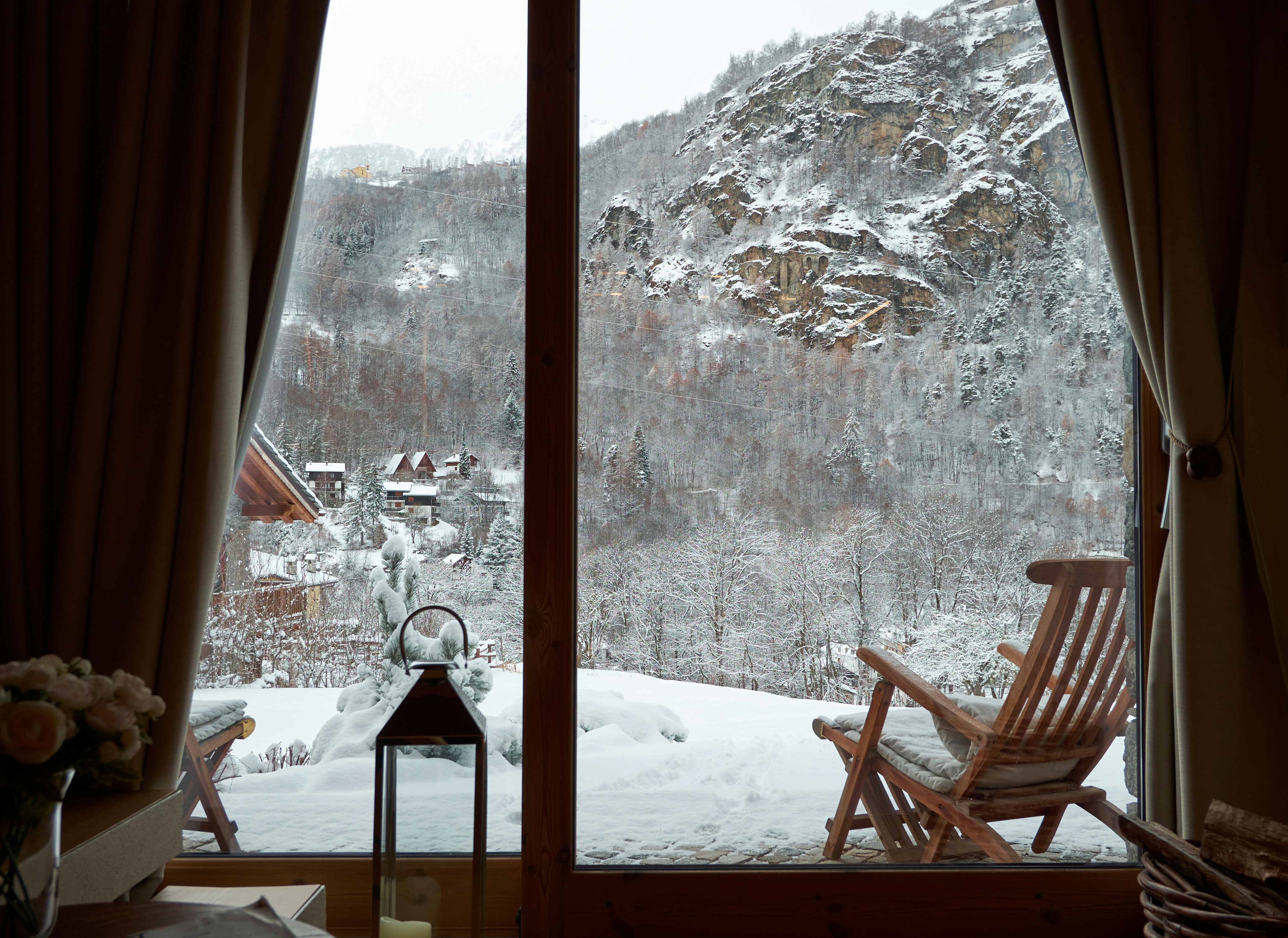 A quiet room with curtains framing a window that looks out on a snowy garden and a wooden lounge chair