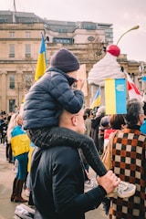 A child wearing a beanie is sitting on an adult's shoulders in a crowd. People are carrying Ukrainian flags and there appears to be a large sculpture of an ice cream cone with a cherry in the background. The event is taking place in an urban environment with historical buildings visible.