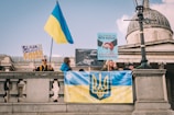 A group of people is gathered on a public square holding banners and flags. The Ukrainian flag is prominently displayed, along with signs advocating for Ukraine. The architecture in the background is classic with stone and dome features, possibly a significant public building.