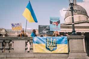 A group of people is gathered on a public square holding banners and flags. The Ukrainian flag is prominently displayed, along with signs advocating for Ukraine. The architecture in the background is classic with stone and dome features, possibly a significant public building.