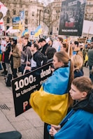 A group of people gathered at a demonstration, many carrying signs and flags. The signs convey messages related to the situation in Ukraine, and some involve imagery of a maternity center in Mariupol. A woman and a child are wrapped in a Ukrainian flag, highlighting the themes of protest and solidarity.