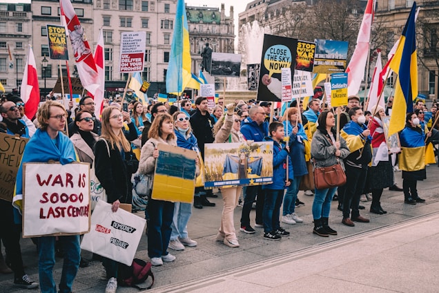 A group of people gathered in a public square holding signs and banners in support of Ukraine. Many participants are wearing Ukrainian flags or colors, and several signs with messages for peace and solidarity are visible. The background shows a cityscape and a fountain.