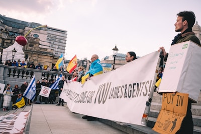 A public gathering with people holding various flags and banners, including Ukrainian and Israeli flags. One person holds a sign with a statement against Putin. The scene takes place on a stone-paved area near an urban architectural setting. The atmosphere is vibrant and charged with political messages.
