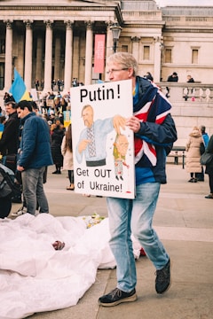 A protestor holding a sign that reads 'Putin! Get OUT of Ukraine!' featuring a cartoonish depiction. The setting appears to be in front of a historical building with columns and a gathered crowd. Several people hold flags and signs, indicating a protest or demonstration.