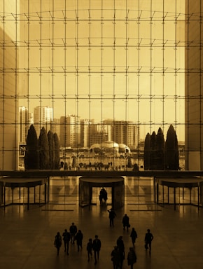 A large indoor space with floor-to-ceiling windows showcasing a view of an urban cityscape. The glass facade is segmented into a grid, while a small group of silhouetted people walk inside, approaching revolving doors. Outside, tall buildings and trees are visible under a sepia tone.