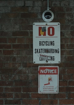 A wall with two warning signs, featuring a video surveillance camera mounted above. The upper sign prohibits activities such as bicycling, skateboarding, and loitering. The lower sign alerts that all activities are monitored by video camera. Both signs are attached to a textured brick wall.