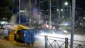 A nighttime urban scene with a street lined by large waste bins, some of which are overflowing with trash. On the left, there is visible steam or smoke rising from one of the bins. In the background, a busy street filled with cars is illuminated by streetlights. Several buildings stand tall in the distance, adorned with billboards and lit windows.