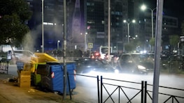 A nighttime urban scene with a street lined by large waste bins, some of which are overflowing with trash. On the left, there is visible steam or smoke rising from one of the bins. In the background, a busy street filled with cars is illuminated by streetlights. Several buildings stand tall in the distance, adorned with billboards and lit windows.