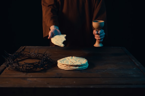 A person in a brown robe is holding a piece of broken bread in one hand and a goblet in the other. On the wooden table in front of the person, there is a stack of flatbreads and a crown of thorns. The background is dark.