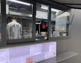 A person is working inside a street food or beverage kiosk, visible through a large service window. The kiosk has a menu in both English and another language, offering various items like teas, smoothies, and sandwiches. Inside, there are several kitchen equipment items and food supplies, suggesting a place for preparing orders.
