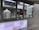 A person is working inside a street food or beverage kiosk, visible through a large service window. The kiosk has a menu in both English and another language, offering various items like teas, smoothies, and sandwiches. Inside, there are several kitchen equipment items and food supplies, suggesting a place for preparing orders.