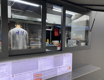 A person is working inside a street food or beverage kiosk, visible through a large service window. The kiosk has a menu in both English and another language, offering various items like teas, smoothies, and sandwiches. Inside, there are several kitchen equipment items and food supplies, suggesting a place for preparing orders.