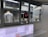 A person is working inside a street food or beverage kiosk, visible through a large service window. The kiosk has a menu in both English and another language, offering various items like teas, smoothies, and sandwiches. Inside, there are several kitchen equipment items and food supplies, suggesting a place for preparing orders.