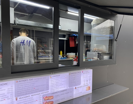 A person is working inside a street food or beverage kiosk, visible through a large service window. The kiosk has a menu in both English and another language, offering various items like teas, smoothies, and sandwiches. Inside, there are several kitchen equipment items and food supplies, suggesting a place for preparing orders.