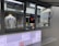 A person is working inside a street food or beverage kiosk, visible through a large service window. The kiosk has a menu in both English and another language, offering various items like teas, smoothies, and sandwiches. Inside, there are several kitchen equipment items and food supplies, suggesting a place for preparing orders.