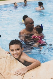 A child in a swimming pool is smiling directly at the camera, holding onto the pool edge. In the background, several people are enjoying the pool, including an adult holding a small child.