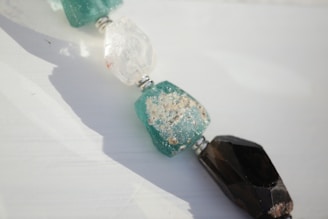 A close-up of three beads, including a translucent white bead, a rough turquoise bead with speckles, and a faceted dark bead. The beads are strung together with small metallic spacers in between. The background is softly lit, giving a natural and peaceful feel.