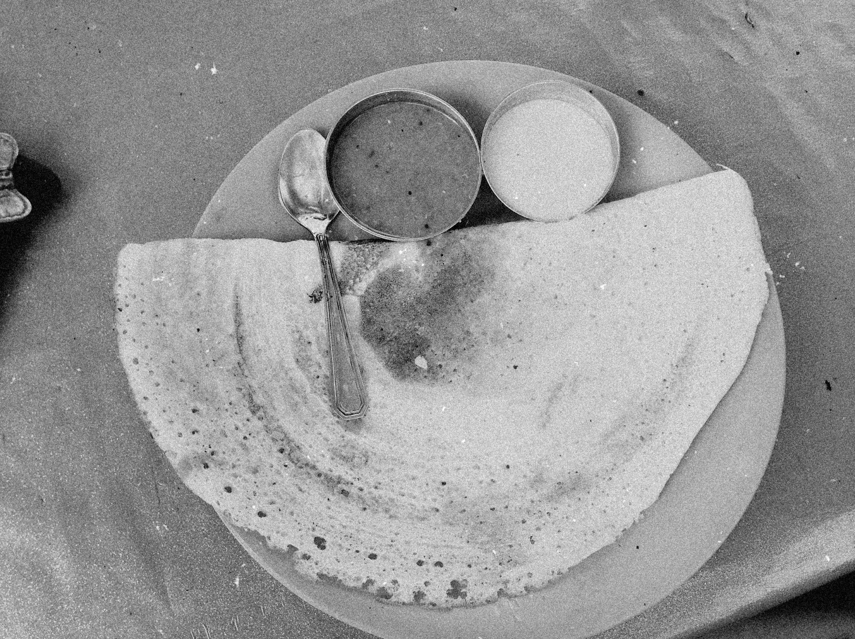 A black-and-white photo of a folded dosa on a plate with small steel bowls of sambar and coconut chutney